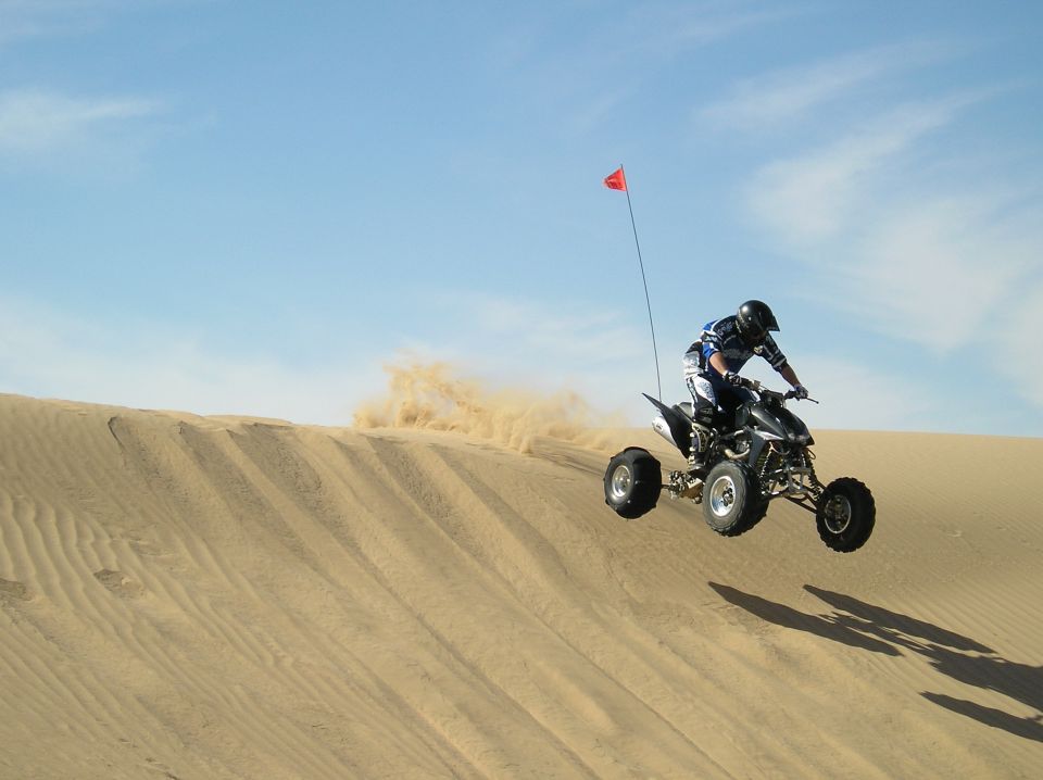 Quad bike mid-air over a sand dune, kicking up dust. Red flag on a pole is visible