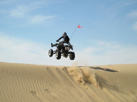 1993 Ford Bronco - A rider on a quad bike soars through the air over a sand dune, kicking up dust.