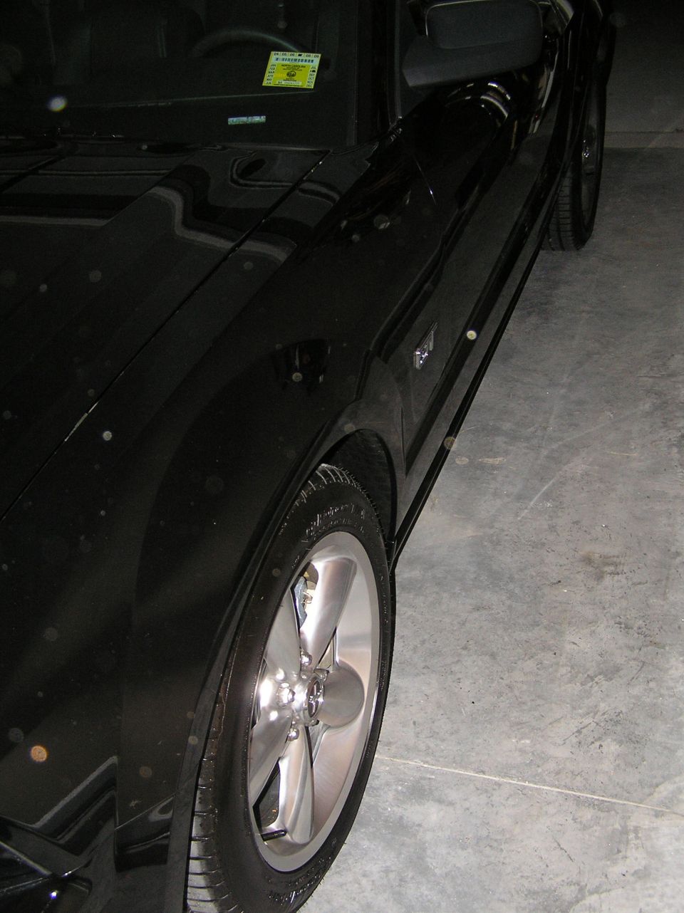 Black Ford Mustang front driver's side with five-spoke alloy wheel in a garage