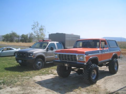 1979 Ford Bronco - An orange and blue Ford Bronco sits lifted on large tires, parked next to a silver truck under a clear blue sky.