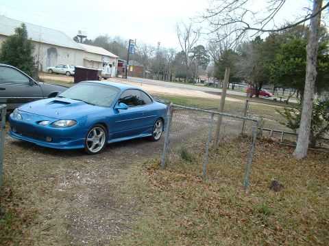 2000 Ford Escort - A modified blue coupe sits in a gravel lot next to a chain-link fence.