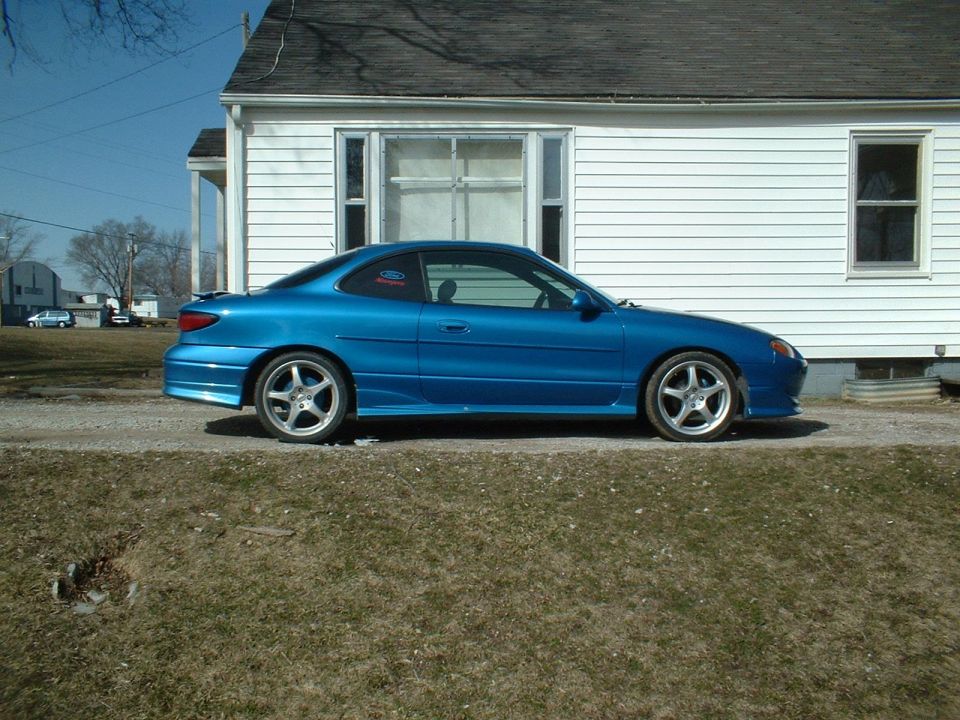 Blue 2000 Ford Escort coupe parked on gravel in front of a white house
