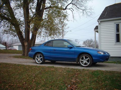 2000 Ford Escort - A blue coupe sits parked on a dirt driveway, with a large tree to its left and a white house to its right.