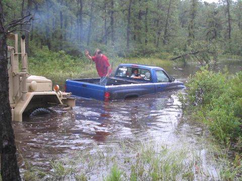 1995 Dodge 1500 - A blue pickup truck is stuck in the water, with a person waving from its bed while a military truck is nearby.