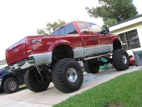 1974 Ford F-100 - A lifted red pickup truck with prominent off-road tires is parked on a driveway, with another vehicle partially visible to the left.