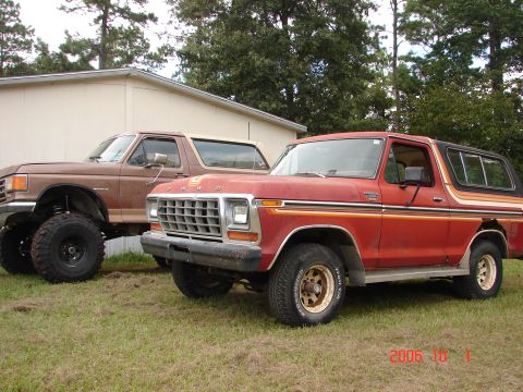 1987 Ford Bronco - Two full-size Ford Broncos parked on grass beside a cream mobile home: in front, a red and tan late-1970s Bronco Ranger XLT with FORD block…