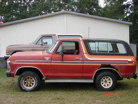 1987 Ford Bronco - A red Ford Bronco with a black top sits in front of a building, with a brown pickup truck behind it.