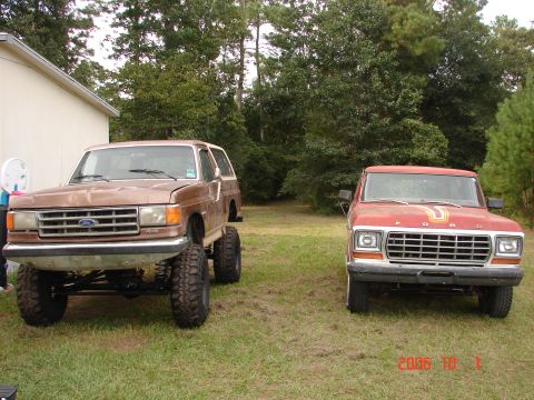 1987 Ford Bronco - Two Ford 4x4s parked side by side on a grassy lot at the edge of pine woods: a lifted brown Bronco with a tan top, no front bumper and expo…