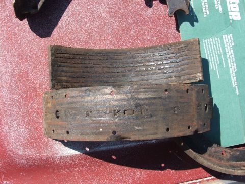 1966 Ford Ranchero - Two worn drum brake shoes laid on a red tabletop, the upper one showing grooved, heat-glazed friction lining and the lower one flipped to s…