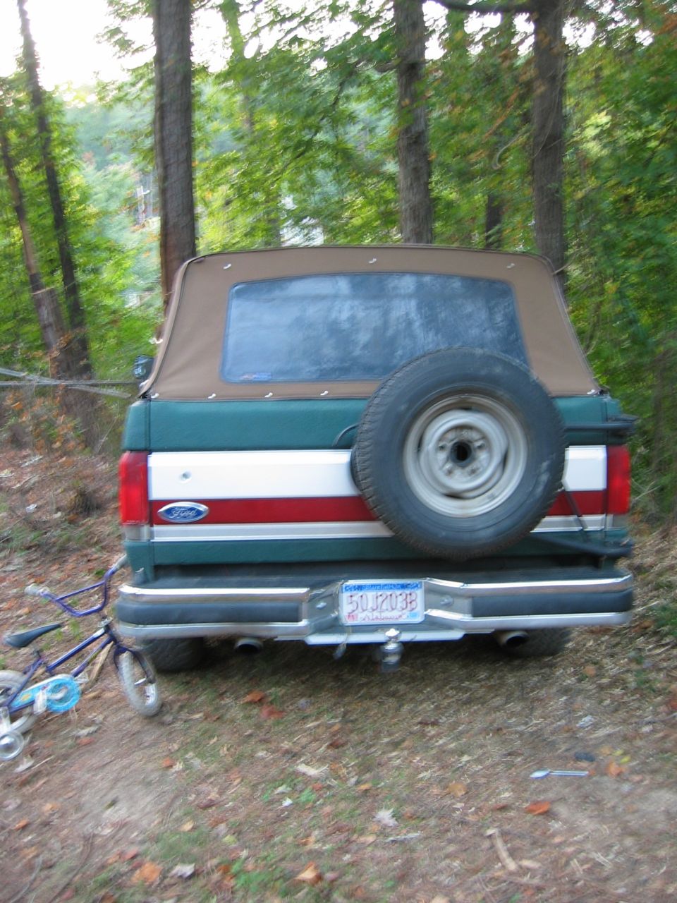 1990 Ford Bronco rear view, green and white with red stripe, soft top, spare tire on back. Bicycle to the left