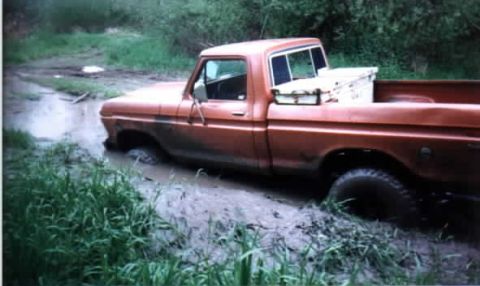 1976 Ford F-100 - This orange pickup truck is stuck deep in the mud, with its front end submerged in water and muck.