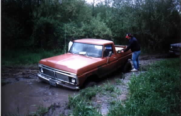 Orange 1970s Ford pickup stuck to the rockers in a mud hole with a person standing alongside