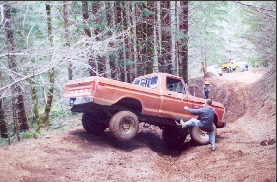 Orange 1970s Ford 4x4 pickup flexing with a front wheel lifted on a steep dirt trail