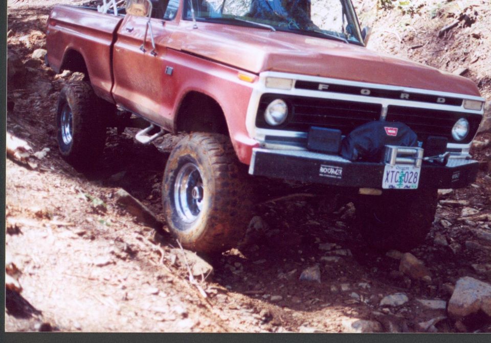 Orange-red 1970s Ford 4x4 pickup with Warn winch flexing over a rocky trail, front three-quarter view