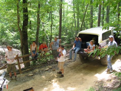 1992 Ford Mustang - Several people are gathered around a parked SUV in a wooded area, some sitting on a fence and others on the vehicle's tailgate.