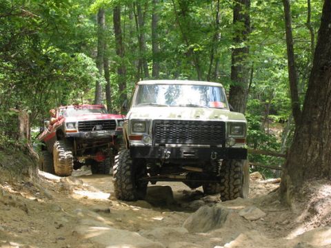 1992 Ford Mustang - Two modified trucks navigate a challenging rocky trail through a dense forest.