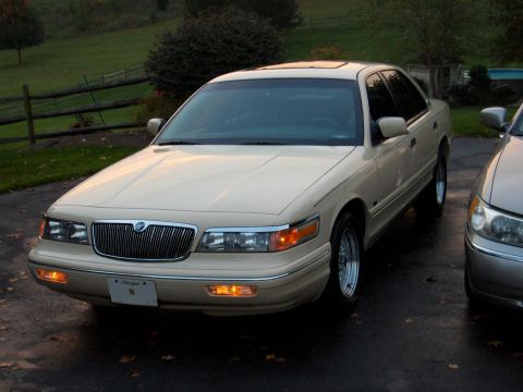 1996 Mercury Grand Marquis - A tan sedan with a chrome grille is parked on a wet surface next to another vehicle.
