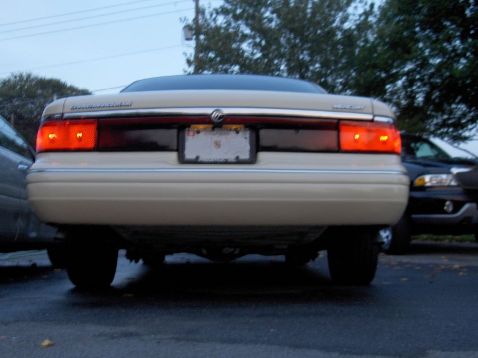 Rear of tan Mercury sedan with lit brake lights, low angle in parking lot