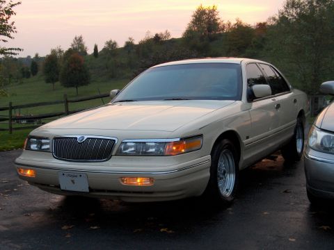 1996 Mercury Grand Marquis - A tan sedan with a chrome grille and aftermarket wheels is parked on wet asphalt near a fence.