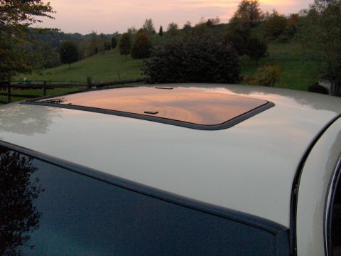 1996 Mercury Grand Marquis - The sunroof of a cream-colored vehicle reflects the sunset sky, set against a backdrop of trees and rolling hills.