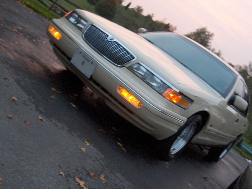 Cream Mercury Grand Marquis sedan, low tilted front 3/4 view, lights on at dusk