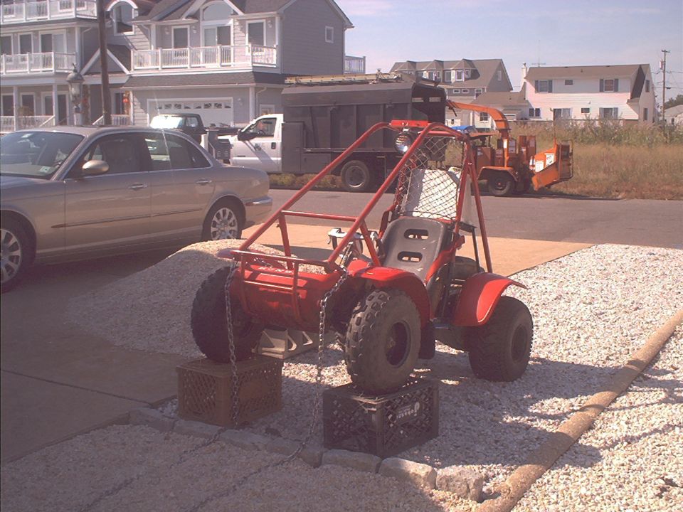 Red off-road buggy on milk crates in gravel driveway with houses behind