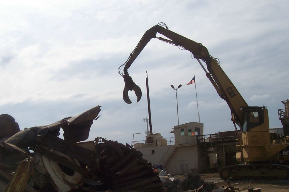 Demolition crane with a claw attachment over a pile of scrap metal, with a ship and American flag in the background