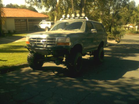 1996 Ford Bronco - A lifted green Ford Bronco is parked on a residential street with a white SUV in the background.