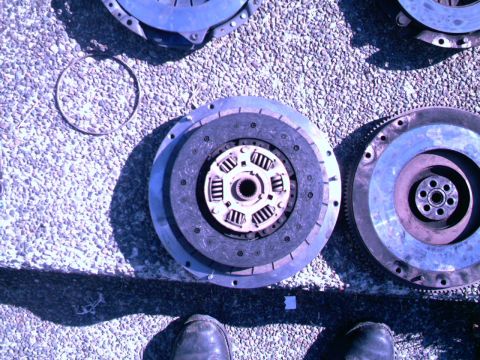 1987 Ford Bronco II - Clutch assembly and flywheel parts are laid out on a gravel surface, with a ring and two feet visible at the bottom.
