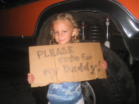 1979 Ford Bronco - A child holds a sign asking for votes in front of a large vehicle's tire and fender.