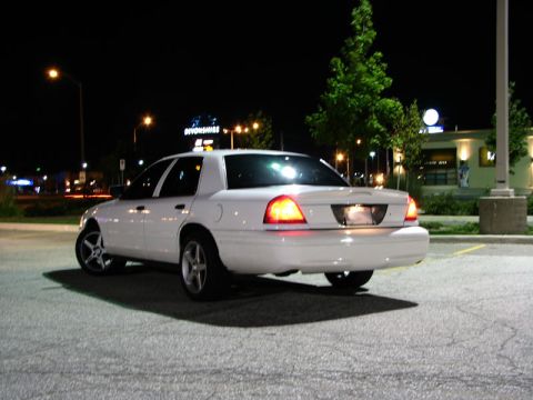 2003 Ford Crown Victoria - Night shot of a white full-size four-door sedan photographed from the rear three-quarter angle in an empty parking lot, tail lights glowing…
