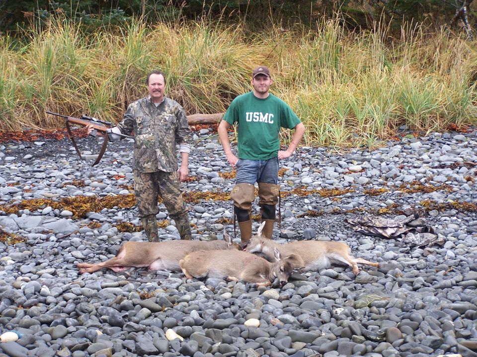 Two hunters stand over four dead deer on a rocky shore, one holding a rifle