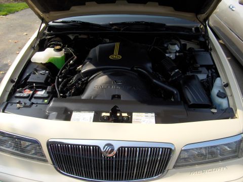 1996 Mercury Grand Marquis - Engine bay view of a cream/tan Mercury sedan with the hood open, showing a black V8 engine cover marked "CAUTION FAN" and yellow accent str…