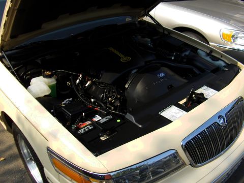 1996 Mercury Grand Marquis - Engine bay of a cream/tan Mercury Grand Marquis with the hood raised, showing a black V8 engine cover, white coolant reservoir, black batte…