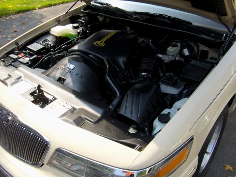 1996 Mercury Grand Marquis - Engine bay of a cream/tan Mercury Grand Marquis with the hood up, showing a black V8 engine cover reading "CAUTION FAN" and "V8," black air…