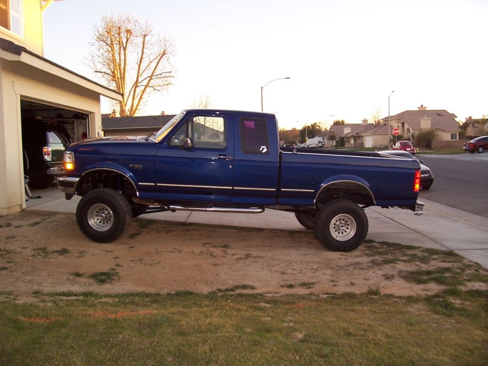 Lifted dark blue Ford F-150 extended cab pickup, side view, parked on dirt driveway by an open garage