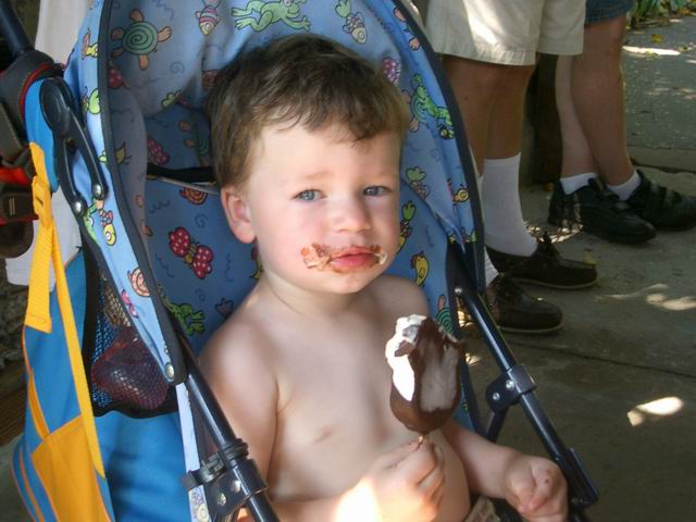 Child with chocolate on face sits in a stroller holding an ice cream bar