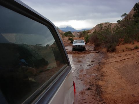 1993 Ford Bronco - Two trucks navigate a wet, muddy path through a desert canyon under an overcast sky.