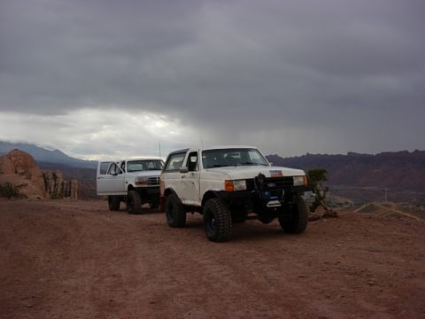 1993 Ford Bronco - Two white Ford Broncos are parked on a dirt road in a mountainous desert landscape under a cloudy sky.