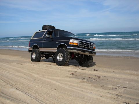 1996 Ford Bronco - A lifted Ford Bronco sits on the sand with the ocean waves in the background.