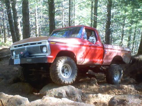 1976 Ford F-100 - A red Ford pickup truck navigates rough terrain in a wooded area, showing its lifted suspension and off-road tires.