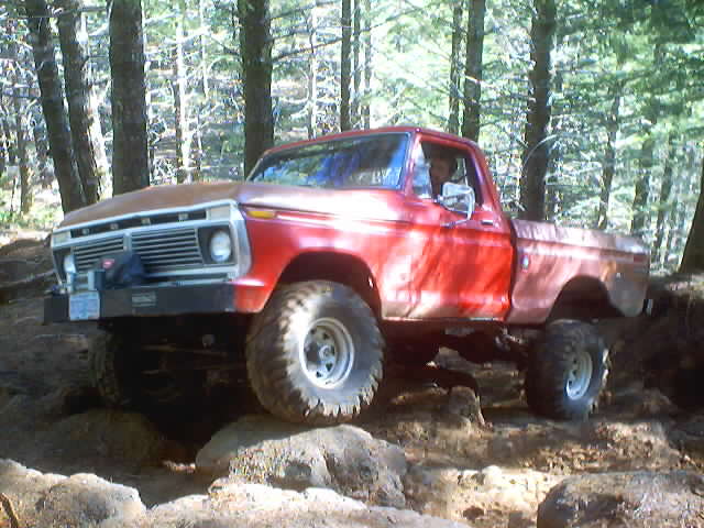 Red Ford pickup truck with a rusty hood, lifted suspension, and large off-road tires, angled to the front