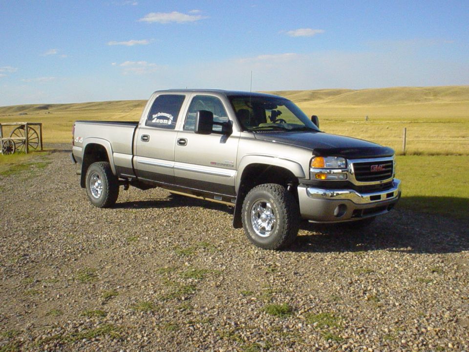 Tan GMC Sierra HD crew cab 4x4 with hood deflector and chrome wheels parked on gravel in open prairie