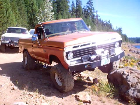 1976 Ford F-100 - A lifted orange-and-white 1970s Ford F-series regular-cab 4x4 photographed from a front three-quarter angle on a rocky pine-lined trail, we…