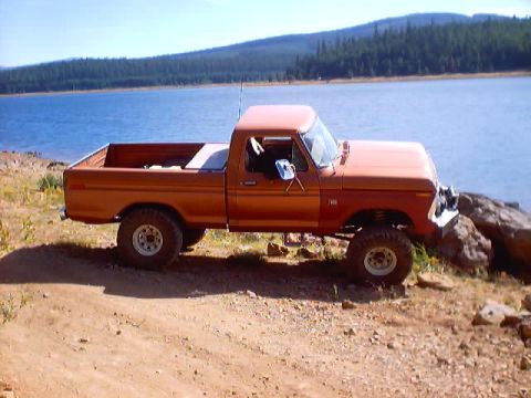 1976 Ford F-100 - Driver-side profile of an orange square-body Ford 4x4 pickup with no doors, parked on a dirt bank above a forest-ringed reservoir. It rides…