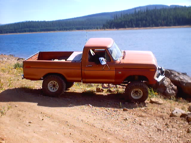 Orange doorless square-body Ford 4x4 pickup parked on a dirt bank above a forested lake