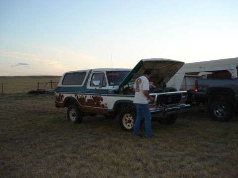 1979 Ford Bronco - Man inspects the engine of a weathered Ford Bronco. A second truck is visible in the background.
