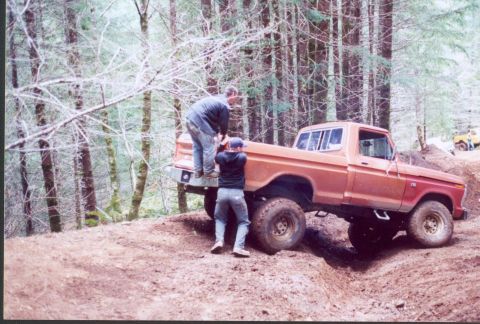 1976 Ford F-100 - A lifted orange 1970s Ford F-series short-bed regular-cab 4x4 sits nose-down on a rutted red-clay trail in a fir forest, viewed from the dr…