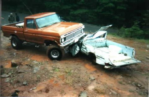 1976 Ford F-100 - An orange 1970s Ford F-series 4x4 pickup sits on a muddy gravel pullout with its front end pushed into a wrecked white pickup bed and body…