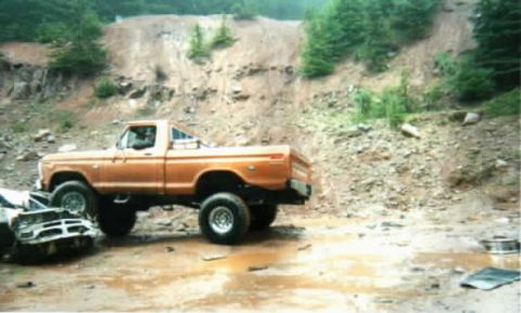 1976 Ford F-100 - This lifted orange pickup truck is parked on muddy terrain next to a damaged vehicle, with a steep, rocky incline behind it.